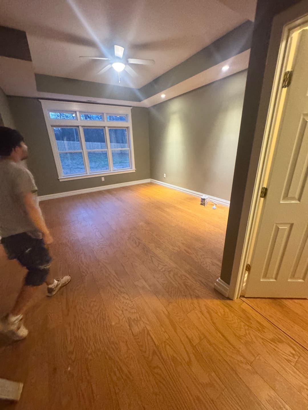 Person standing in an empty room with wooden flooring and green walls, ceiling fan above.