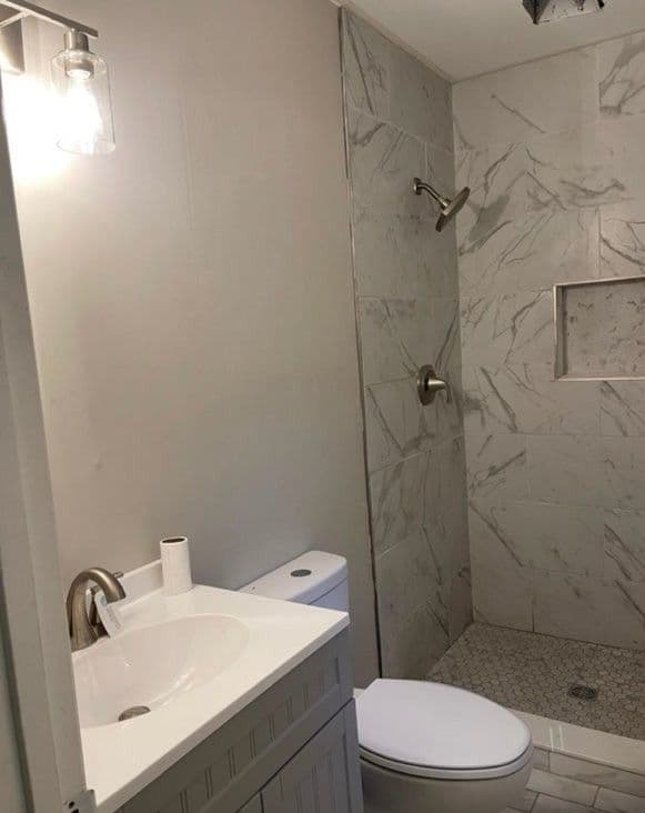 Modern bathroom with marble shower, white vanity, toilet, and stylish lighting fixture.
